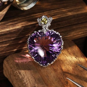 Peut inclure: Un pendentif en améthyste violette en forme de cœur serti dans une monture en argent texturé. Le pendentif présente une petite pierre précieuse ronde jaune sur la bélière supérieure, également en argent texturé, offrant un aspect détaillé.