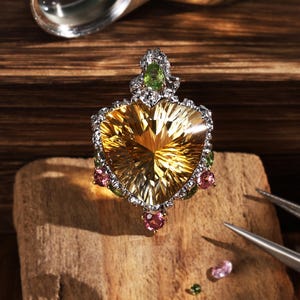 Peut inclure: Un pendentif avec une grande pierre précieuse jaune facettée en forme de cœur comme pièce centrale. La monture est en métal argenté ornée de petites pierres roses et vertes rondes. Le design inclut une pierre verte en forme de poire au sommet.