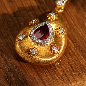 Könnte beinhalten: Ein goldener Anhänger mit einem tropfenförmigen Rubin, der von Diamanten umgeben ist. Der Anhänger ist in einer goldenen Fassung mit einer strukturierten Oberfläche gefasst und mit kleineren Diamanten in einem Sternmuster verziert.