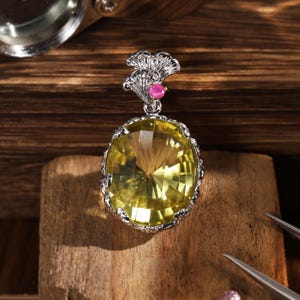 Puede incluir: Un colgante de plata con una gran piedra preciosa amarilla de corte ovalado engastada en un marco de metal texturizado. El pasador tiene forma de hoja de ginkgo y está acentuado con una pequeña piedra preciosa redonda de color rosa vibrante.