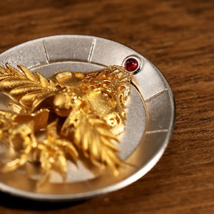 May include: A detailed close-up of a round, silver-colored metal object with a gold-colored, ornate design of a mythical creature. A small, red gemstone is set into the silver border. The object is on a wooden surface.