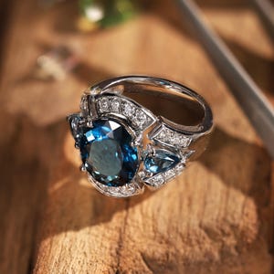 May include: A silver ring featuring a large oval-cut dark blue gemstone as the centerpiece, flanked by two smaller pear-cut blue stones. The band and setting are accented with numerous small, clear round stones and stamped with S925.