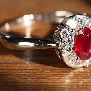 Può includere: Un anello d'argento con un grande rubino ovale circondato da diamanti più piccoli. L'anello è incastonato su una fascia di metallo bianco.