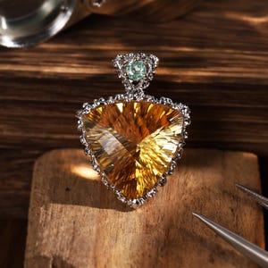 Peut inclure: Un pendentif triangulaire avec une grande pierre précieuse jaune doré à facettes, sertie dans une monture en argent texturé. Une petite pierre précieuse ronde vert clair est fixée sur la bélière supérieure, créant des reflets lumineux.