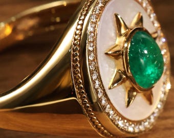 May include: A gold ring with an emerald gemstone at its centre, set within a starburst design. The ring has a white background with a border of small diamonds. The band is polished gold.