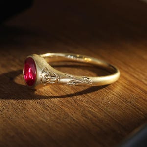 Könnte beinhalten: Ein goldener Ring mit einem roten Edelstein und einem floralen Design. Der Ring liegt auf einer Holzoberfläche.