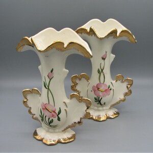 May include: Two off-white ceramic vases with gold trim and pink floral designs. The vases have a unique shape with a ruffled top and decorative side elements. The floral design includes pink flowers and green leaves.