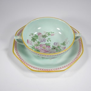May include: A light blue soup bowl with two handles and a floral design. The bowl is sitting on a matching saucer with a scalloped edge.