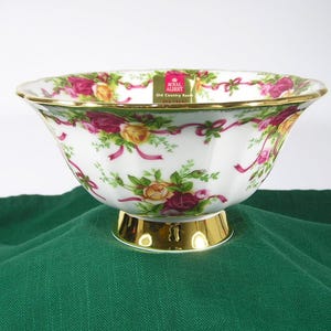 May include: A white porcelain bowl with a gold rim and a floral pattern featuring roses, ribbons, and greenery. The bowl is on a gold pedestal and has the words "Royal Albert Old Country Roses" printed on the side.