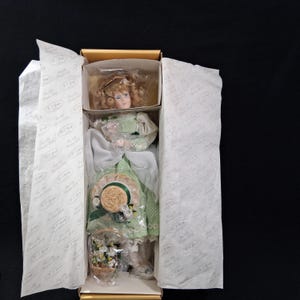 May include: A porcelain doll in a box, featuring a green polka-dot dress, a matching hat, and a basket of flowers. The doll has blonde curly hair and is packaged with tissue paper.