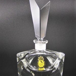 May include: A clear glass bottle with a faceted design and a geometric stopper. The bottle has a gold label with the text "IMPERIAL CRYSTAL HANDMADE IN GERMANY".