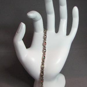 May include: A silver bracelet with oval-shaped links, each containing a small, colorful gemstone. The gemstones are in shades of red, green, and clear. The bracelet is displayed on a white hand-shaped jewelry holder against a gray background.