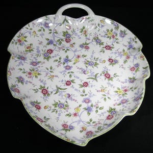 May include: White ceramic serving platter with a floral pattern in shades of pink, purple, yellow, and blue. The platter has a leaf-like shape with a handle and a scalloped edge. The floral design covers the entire surface.