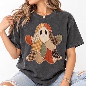 May include: A charcoal gray short-sleeve t-shirt featuring a large ghost-shaped graphic made of various fabric patches. The ghost design includes patterns like gingham, plaid, and floral prints in shades of orange, brown, cream, and olive green.