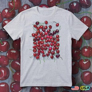 May include: A light gray short-sleeve t-shirt featuring a central graphic of numerous red cherries with green stems. Behind the cherries, there is faint, illegible cursive script text printed in a light gray color across the chest area.