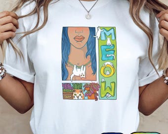 Puede incluir: Camiseta blanca con un gráfico colorido que presenta a una mujer con cabello azul, un gato y la palabra "MEOW". El diseño incluye un gato colgando de un collar, un gato en una maceta y otras ilustraciones de gatos. La camiseta tiene cuello redondo.