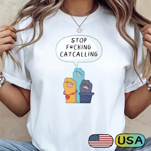May include: White t-shirt with the text "STOP F*CKING CATCALLING" in a speech bubble above three cartoon cat paws. The paws are yellow, blue, and purple, each with unique details. A USA flag graphic is at the bottom.