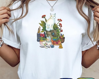 Puede incluir: Camiseta blanca con una ilustración caprichosa de un gato blanco rodeado de plantas, útiles de arte y una pequeña estatua de Buda. El gato está posado sobre un recipiente de madera. El diseño incluye flores rojas y un gato durmiente. La camiseta tiene cuello redondo.