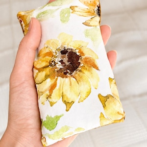 Op de afbeelding: Een rechthoekig stoffen zakje met een aquarelprint van gele zonnebloemen met bruine harten en groene bladeren op een witte achtergrond. Het item heeft een zacht, gevuld uiterlijk en wordt in een handpalm getoond om het bloemmotief te tonen.