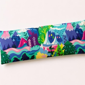 May include: A rectangular pillow with a mountain landscape design. The pillow features a vibrant pattern of purple, pink, green, and blue mountains, trees, and flowing water. The pillow is approximately 30 cm long.
