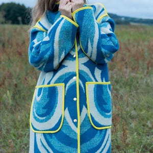 May include: A long coat with a blue and white abstract pattern. The coat has yellow trim around the edges, pockets, and collar. It is buttoned up and appears to be made of a thick, woven fabric. The coat is worn outdoors in a field.