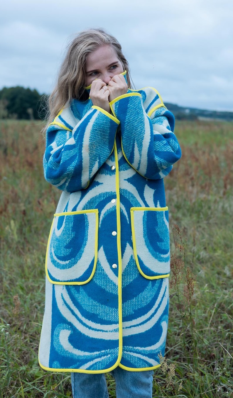 May include: A long coat with a blue and white abstract pattern. The coat has yellow trim around the edges, pockets, and collar. It is buttoned up and appears to be made of a thick, woven fabric. The coat is worn outdoors in a field.
