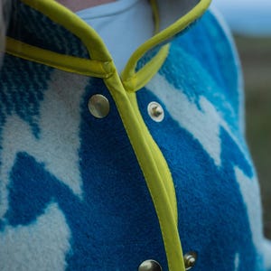 May include: Close-up of a blue and white patterned jacket with a yellow trim and gold buttons. The jacket has a unique design and appears to be made of a soft, textured fabric. The collar is also trimmed with yellow.