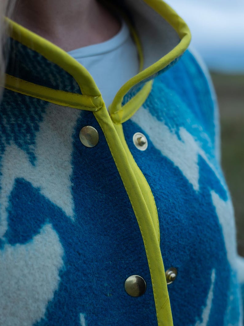May include: Close-up of a blue and white patterned jacket with a yellow trim and gold buttons. The jacket has a unique design and appears to be made of a soft, textured fabric. The collar is also trimmed with yellow.
