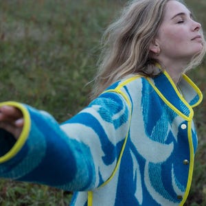 May include: A blue and white patterned jacket with yellow trim. The jacket has a button-up front and a unique wave-like design. The sleeves are long and the collar is yellow.
