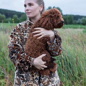 May include: A woman wearing a brown patterned jacket and blue jeans holds a brown curly-haired dog in a field. The jacket has a detailed floral and geometric design. The dog's fur is a rich, reddish-brown color.