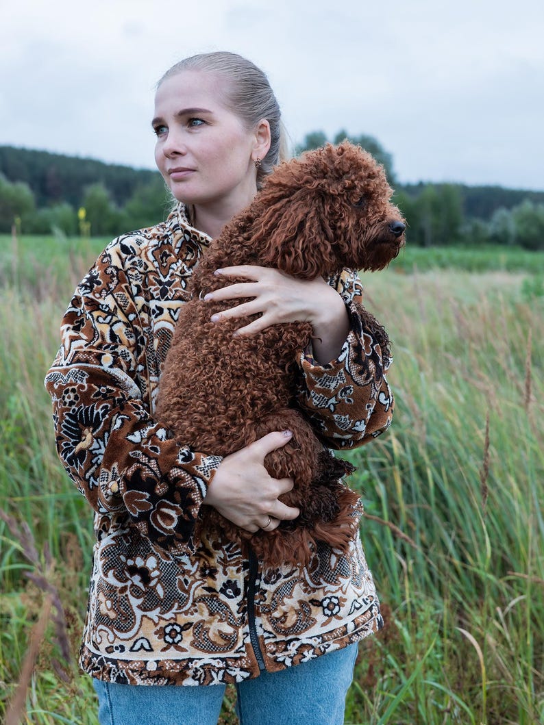 May include: A woman wearing a brown patterned jacket and blue jeans holds a brown curly-haired dog in a field. The jacket has a detailed floral and geometric design. The dog's fur is a rich, reddish-brown color.