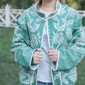 May include: A teal and white floral patterned jacket with a high collar and silver snap button closures. The jacket features long sleeves and white trim along the front opening, collar, and cuffs, worn over a plain white shirt.