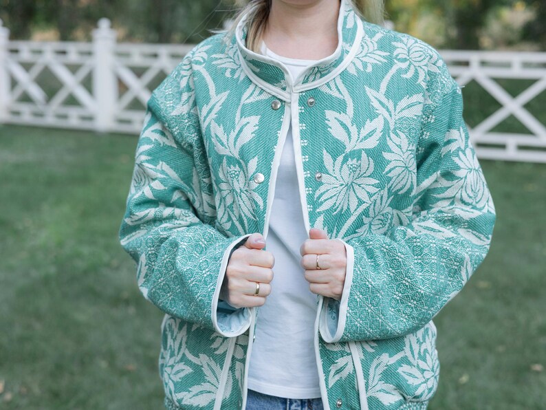 May include: A teal and white floral patterned jacket with a high collar and silver snap button closures. The jacket features long sleeves and white trim along the front opening, collar, and cuffs, worn over a plain white shirt.