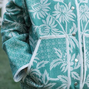 May include: A teal and white jacket featuring a prominent floral pattern with a textured weave. The garment includes a white piped trim along the front opening and pockets, secured with silver-toned metal snap buttons down the center front.