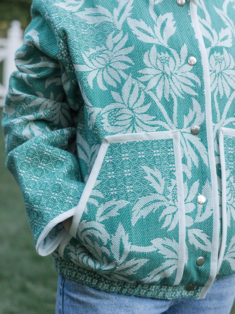 May include: A teal and white jacket featuring a prominent floral pattern with a textured weave. The garment includes a white piped trim along the front opening and pockets, secured with silver-toned metal snap buttons down the center front.