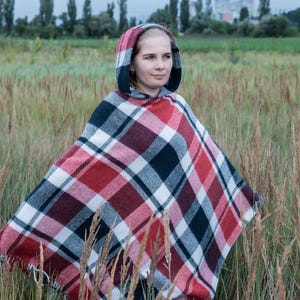 May include: A hooded poncho in a red, black, and white plaid pattern. The poncho is worn outdoors in a field of tall grass. The plaid pattern is a classic design, suitable for cool weather.