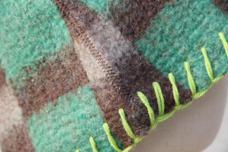 Puede incluir: Primer plano de una manta de lana con un estampado de cuadros en tonos verdes, marrones y grises. La manta tiene un borde cosido con hilo verde brillante. La textura de la lana es visible.