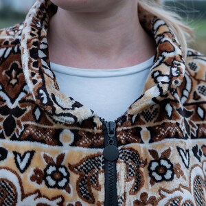 May include: A patterned zip-up jacket in shades of brown, tan, and cream. The jacket features a detailed floral and geometric design. The black zipper runs down the center. The collar and cuffs are also patterned. A white shirt is visible underneath.