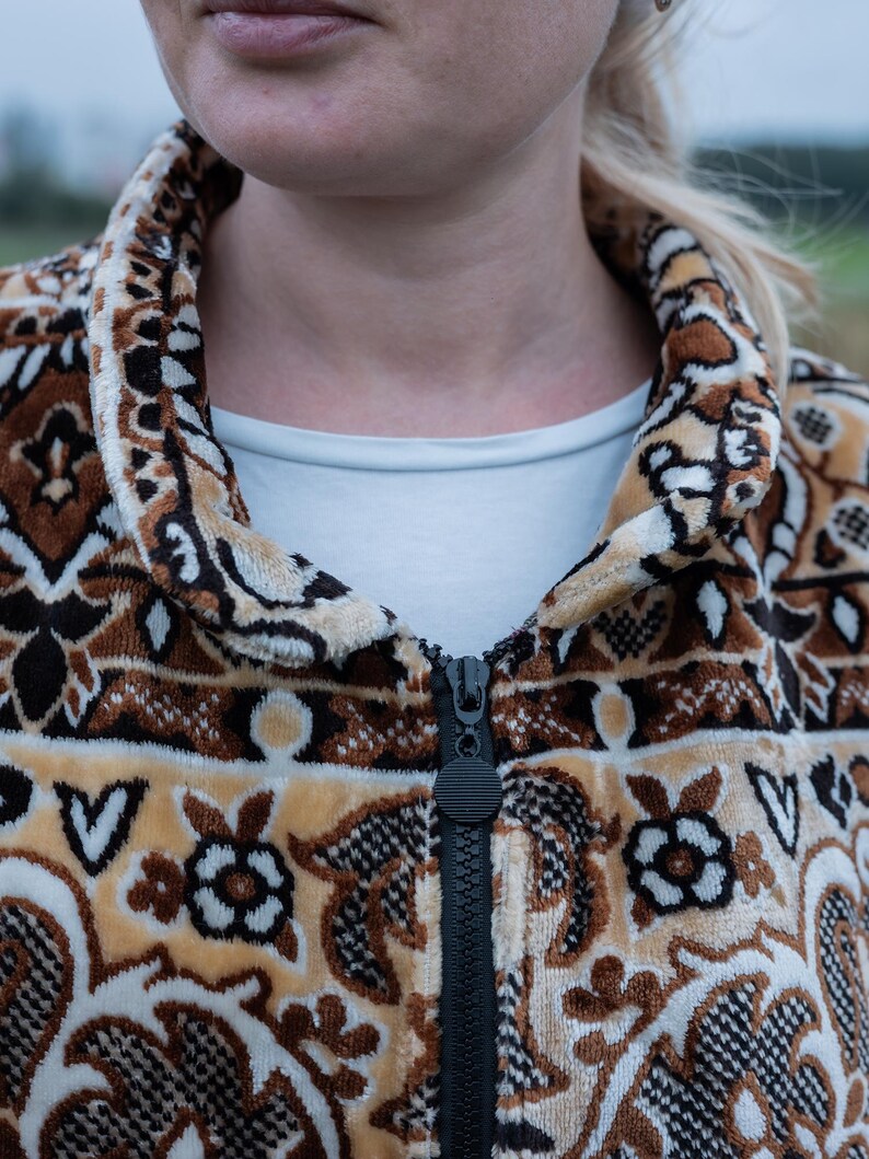 May include: A patterned zip-up jacket in shades of brown, tan, and cream. The jacket features a detailed floral and geometric design. The black zipper runs down the center. The collar and cuffs are also patterned. A white shirt is visible underneath.
