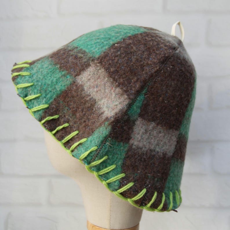Puede incluir: Un sombrero de lana a cuadros en tonos verdes, marrones y grises, con costuras verde brillante a lo largo del borde. El sombrero tiene un lazo en la parte superior y se muestra en una cabeza de maniqu&iacute;.