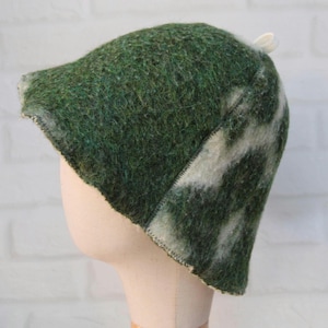 Puede incluir: Un sombrero de lana afieltrada con un patrón jaspeado de fibras verde bosque profundo y blanco roto. El sombrero tiene una copa redondeada, un ala corta y un pequeño lazo blanco unido a la parte superior. La textura es gruesa, difusa y densa.