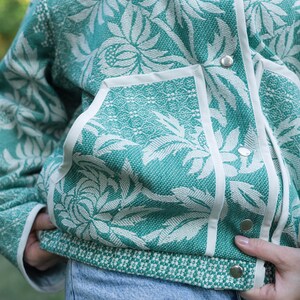 May include: A teal and white floral patterned jacket featuring a textured woven fabric, a front snap button closure with silver-toned hardware, white trim along the pockets and placket, and a contrasting geometric pattern at the bottom hem.