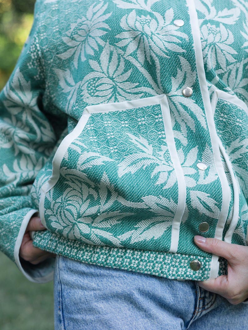 May include: A teal and white floral patterned jacket featuring a textured woven fabric, a front snap button closure with silver-toned hardware, white trim along the pockets and placket, and a contrasting geometric pattern at the bottom hem.