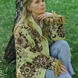 May include: A light green jacket featuring a dark brown and tan floral pattern, long sleeves with fringed cuffs, a high collar, and a matching fringed hemline. The garment is worn with light blue denim jeans, creating a textured look.