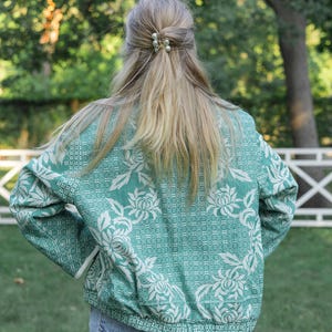 May include: A long-sleeved jacket in a vibrant teal green featuring a white floral pattern with intricate woven textures. The jacket has a ribbed hem and cuffs, a high collar, and a full-length front zipper. It is paired with blue denim jeans.
