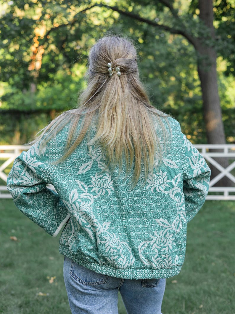 May include: A long-sleeved jacket in a vibrant teal green featuring a white floral pattern with intricate woven textures. The jacket has a ribbed hem and cuffs, a high collar, and a full-length front zipper. It is paired with blue denim jeans.