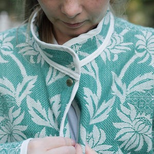 May include: A teal and white jacket featuring a woven floral pattern throughout. The garment has a high collar with white trim along the edges and a single visible metallic snap button closure at the center front of the neckline.