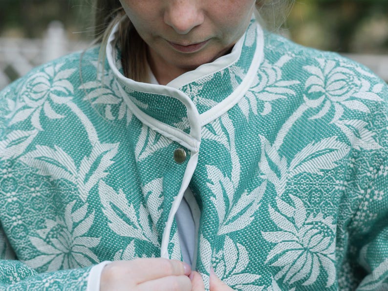 May include: A teal and white jacket featuring a woven floral pattern throughout. The garment has a high collar with white trim along the edges and a single visible metallic snap button closure at the center front of the neckline.