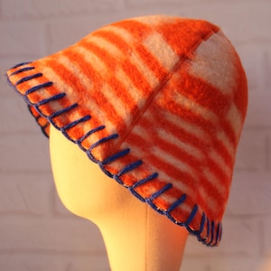 May include: A striped bucket hat in orange and white, with blue stitching around the brim. The hat is made of a soft, textured material. The hat is displayed on a mannequin head.