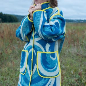 May include: A long coat with a blue and white swirl pattern and yellow trim. The coat has two front pockets and a button-up closure. The person is wearing the coat in a field.